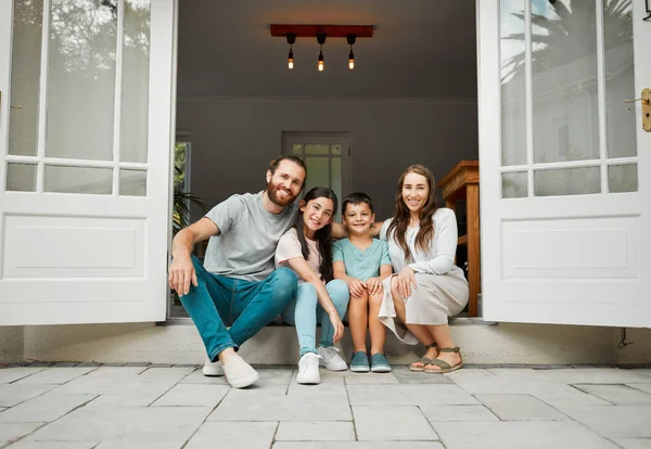 Portrait of a happy young caucasian family of four sitting at their front door smiling and looking at the camera. Two parents sitting with their little son and daughter in front of their house.