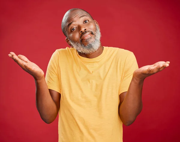 Mature african american man with a beard shrugging his shoulders and making a gesture with his hands standing against a red studio background. Uncertain, decision, choice.