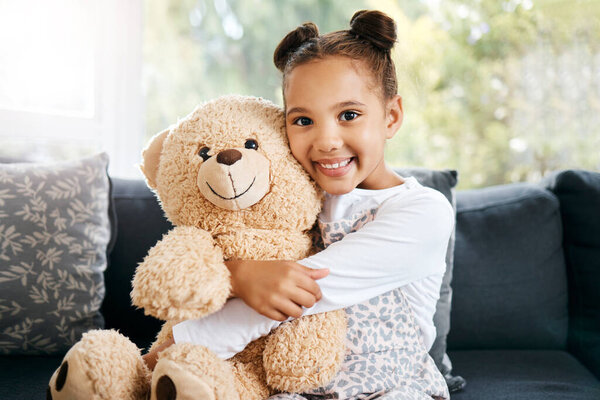 Portrait of an adorable little hispanic girl holding a teddy bear at home. Cute mixed race girl playing with her toy while sitting on the couch in the lounge.