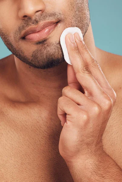 Closeup of one mixed race man wiping a round cotton swab on his face while grooming against a blue studio background. Handsome guy cleaning and exfoliating his face for a healthy complexion and clear.