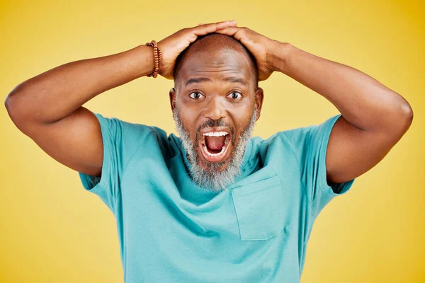 Mature African american man with grey beard standing with his hands on his head and looking shocked or surprised. Man with astonished facial expression while standing against yellow studio background.