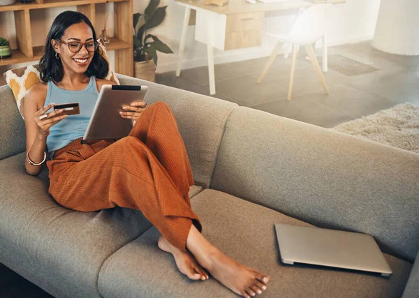 Full length smiling mixed race woman using credit card for ecommerce on digital tablet at home. Happy hispanic sitting alone on sofa, using technology for ebanking. Relaxing, ordering, buying online.