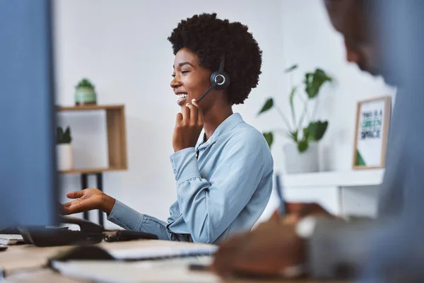 Happy young african american call centre telemarketing agent talking on a headset while working on computer in an office. Confident friendly female consultant operating helpdesk for customer service .