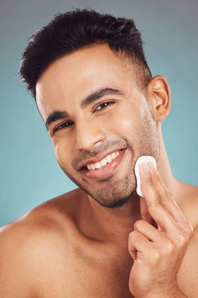 Portrait of one smiling young indian man wiping a round cotton swab on his face while grooming against a blue studio background. Handsome mixed race guy cleaning and exfoliating his face for a health.