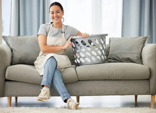 Portrait of a beautiful young mixed race woman holding a laundry basket while cleaning her apartment. One happy Asian woman smiling while folding laundry at home.