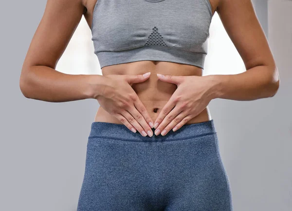 Closeup of one fit caucasian woman framing hands around her slim belly to show weight loss while exercising against a grey background. Female athlete with toned body caring for gut digestion and well.