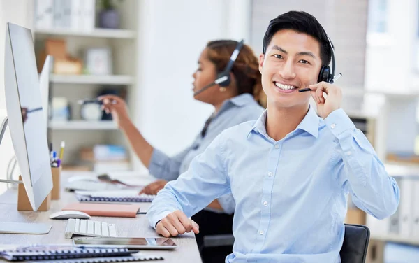 Portrait of asian call centre agent talking on headset while working on computer in office. Confident and smiling businessman consulting and operating a helpdesk for customer sales and service suppor.