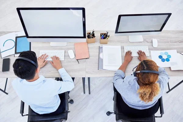 Overhead view of two unknown business people working on computers on a desk in a office. Two colleagues wearing headsets working in a call center.