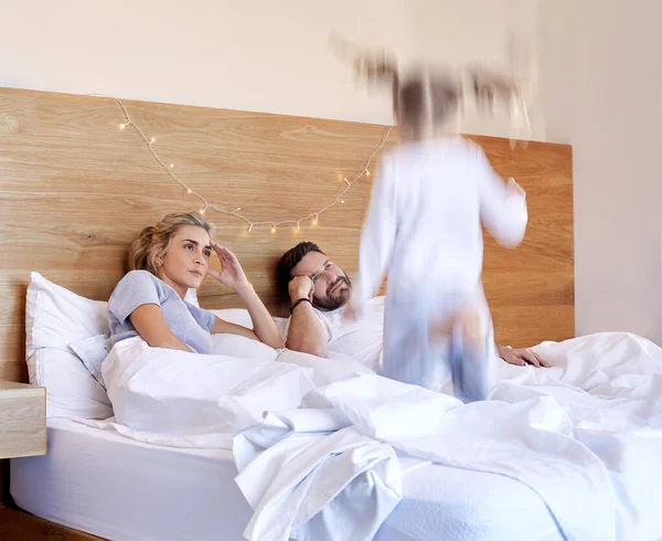 Exhausted parents struggling to put hyperactive toddler child to bed. Tired mom and dad annoyed about noisy active daughter jumping on their bed.