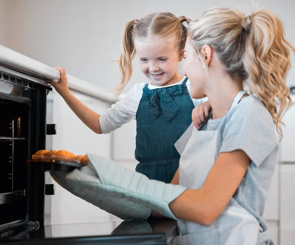 Mother holding tray of muffins. Happy woman taking tray of fresh, baked muffins from the oven. Little girl baking with her mother. Mother and daughter enjoying a tray of dessert muffins.