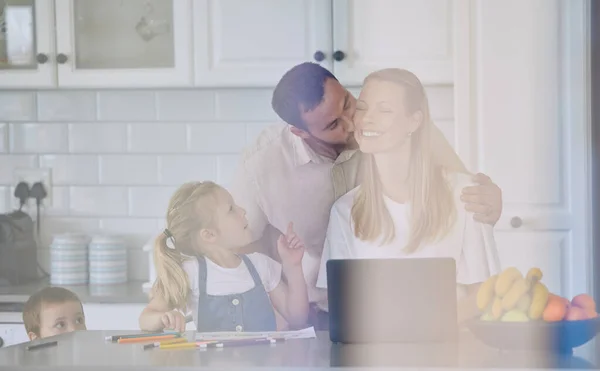 husband kissing wife on the cheek. Family relaxing in the kitchen. Woman working from home on laptop. Mother teleworking with children. Happy family at home together