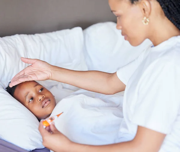 African american woman taking temperature of her son. Mother using thermometer to measure sons temperature. Sick little boy lying in bed. Worried mother feeling sons fever