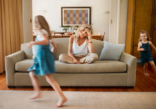 Young unhappy caucasian mother suffering from a headache with her daughters running and playing in the lounge at home. Little siblings being playful together while their mom is stressed.sitting on th.
