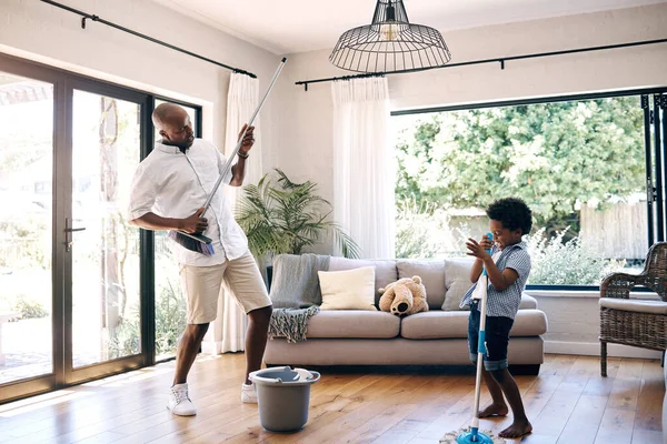 Mature african american dad and his young little son pretending to play the guitar by using a broom in a lounge at home. Black man and his boy having fun while cleaning their home together.