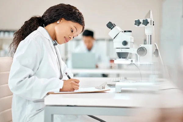彼女の書類を全て管理下に置く 若い科学者が研究室で顕微鏡を使いながらメモを書き — ストック写真