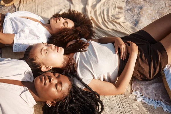 Young diverse female friends laying on the beach together resting with their eyes closed.