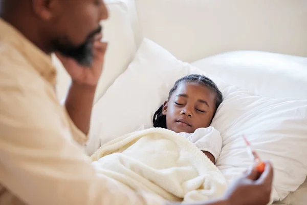 Sick african american girl lying asleep in bed at home. Worried father calling a doctor to help with his unwell daughter after checking her high body temperature with thermometer for symptoms of feve.