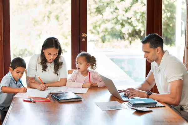 Young mixed race mother helping her children with homework while their father works on a laptop at a table in the lounge. Little siblings drawing together. Hispanic man typing emails.
