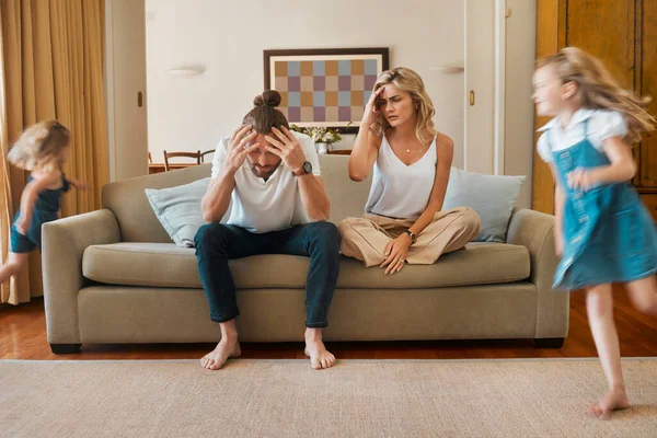Young caucasian mother and father suffering a headache with their daughters running in the lounge at home. Little sisters being playful while their parents are stressed. Woman and man upset sitting o.