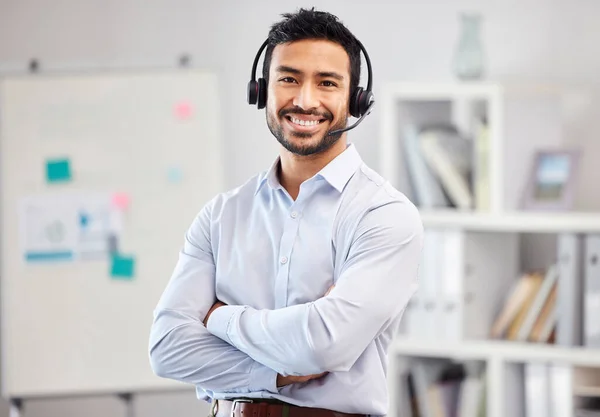 Young happy mixed race call center agent standing with his arms crossed in an office at work. One hispanic customer service agent wearing a headset and answering calls while standing in an office.