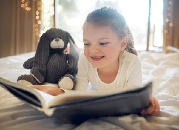 Happy smiling little girl lying on a bed at home and reading a childrens book to her toy teddy. Cute child relaxing and playfully reading a story to her bunny, being creative and playing pretend.