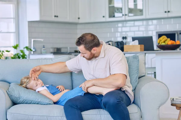 Caucasian father using his hand to feel the high body temperature on his sick little daughters forehead for symptoms of fever, flu or covid. Worried parent caring for ill child lying on sofa at home.