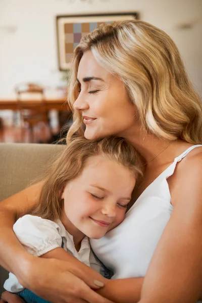 Young loving caucasian mother hugging her little daughter sitting on the couch together at home. Carefree girl embracing her mom resting on the couch.