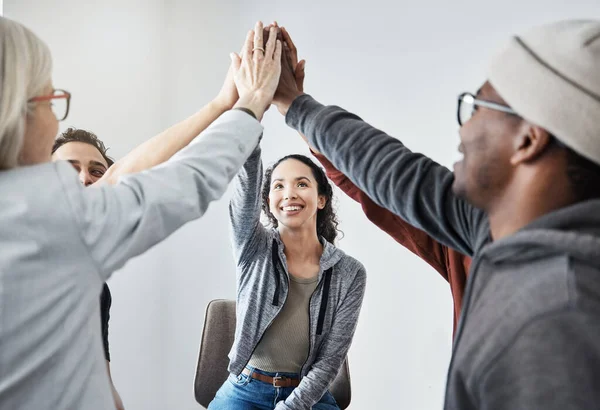 Happy diverse group of businesspeople smiling giving each other a high five sitting in a meeting in an office at work. Happy women and men joining their hands for motivation while working together.