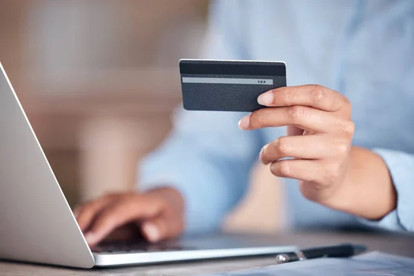 Unrecognizable businessperson holding a credit card in their hand while working on a laptop at a desk at work. One unrecognizable shopping online using their laptop and paying with a card at work.