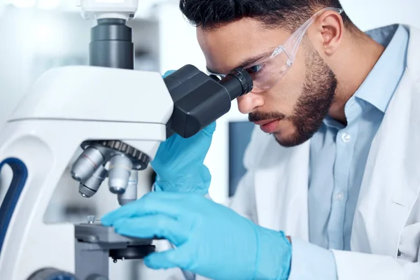 One handsome young mixed race man with a dark beard wearing gloves and a labcoat and looking at medical samples on a microscope in her lab. A male scientist of indian ethnicity wearing safety goggles.