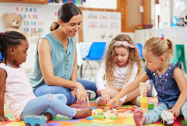 老师和孩子们在幼儿园或学前班做活动时看上去很开心 微笑的老师坐在一个五颜六色的垫子上 和一群不同的幼儿一起玩积木游戏 — 图库照片
