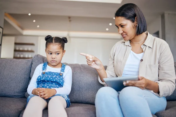 Mixed race woman pointing to scold and discipline her little daughter about internet use on digital tablet at home. naughty girl looking sad while mother punishes her for playing too many games onlin.