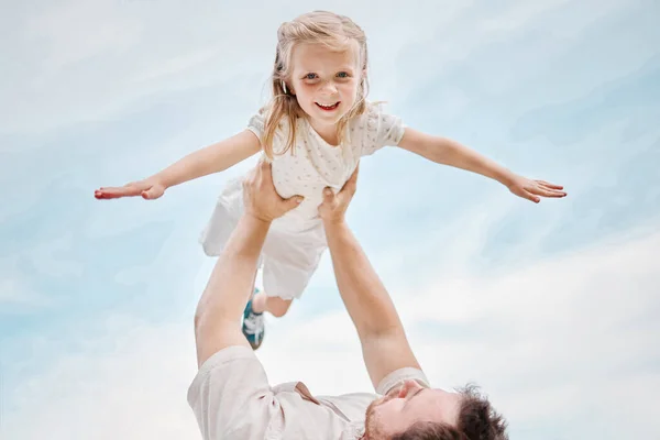 Low angle portrait of an adorable little caucasian girl being carried by her fun playful father while outside in a garden. Smiling cute daughter bonding with her dad in the backyard. Playing aeroplan.