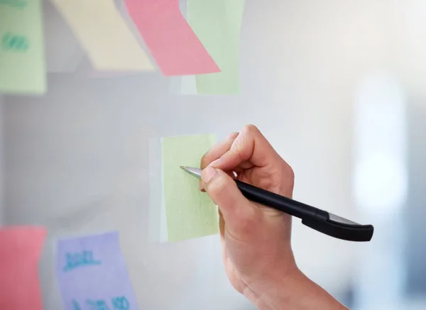 Closeup of unknown business woman writing on sticky notes on a transparent board to brainstorm in an office. Caucasian professional sticking a note on visual aid while planning a strategy with ideas.