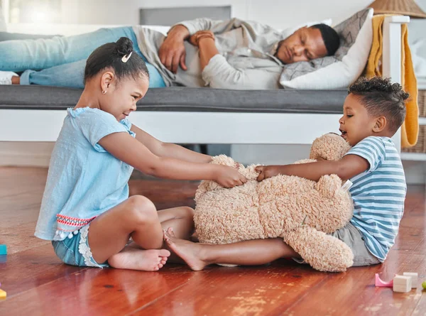 While you were sleeping. two siblings fighting over a teddy on the floor while their dad sleeps on the couch at home