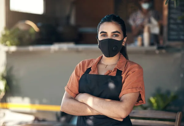 Following Covid rules and regulations. a young woman standing outside her restaurant with her arms folded while wearing a face mask