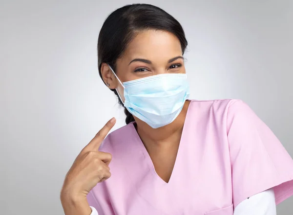 See, its not so bad. Cropped portrait of an attractive young female healthcare worker wearing and pointing towards a mask while standing in studio against a grey background