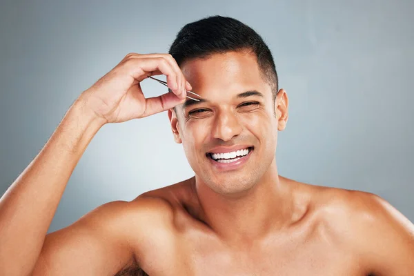 Portrait of a man tweezing his eyebrow with a tweezer in studio with gray background. Happy, smile and young guy plucking brow for hair removal for grooming, cleaning and hygiene routine or self care.