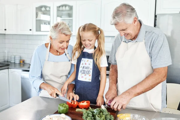 Grandparents, girl child in kitchen and cooking healthy food with vegetables on cutting board for happy family lunch at home. Natural, organic nutrition and clean diet for senior people in retirement.