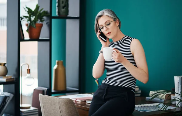Having some coffee while discussing business. a businesswoman having coffee while talking on her cellphone in her office