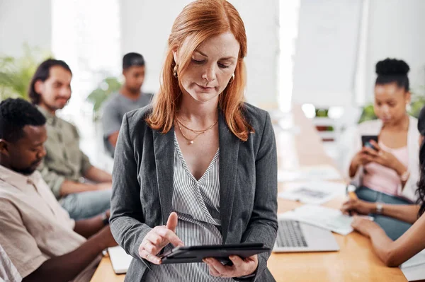 Navigating her way down another path to success. a mature businesswoman using a digital tablet in an office with her colleagues in the background