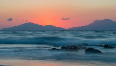 Güneş dağların ardında batarken, altın saatinde uzun bir deniz manzarası. Deniz, kayaların ve kumsalın üzerinden geçerken ruhani görünüyor..