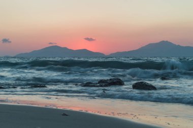 Güneş dağların ardında batarken, altın saatinde uzun bir deniz manzarası. Deniz, kayaların ve kumsalın üzerinden geçerken ruhani görünüyor..