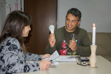 Gülümseyen karı koca elektrik faturalarını tartışırken masada oturuyorlar. Adam kadına, daha fazla tasarruf için eski ampulleri LED ampullerle değiştirmeyi teklif ediyor.