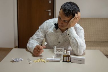 Worried man sitting and leaning on the table at home with anxiolytics and antidepressants in front while holding a test tube in anus. Side effects of psychiatric drugs on male ejaculation