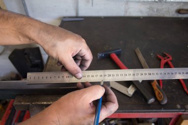 Dirty hands of a blacksmith taking measurements with ruler and pencil on a piece of iron to be cut and worked. Background with tools of the trade