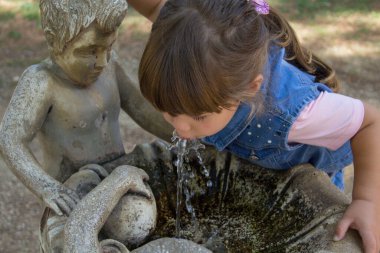 Image of an adorable little girl drinking water from a decorated fountain in a public park. Games and outdoor life with the children during the holidays.
