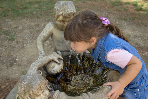 Image of an adorable little girl drinking water from a decorated fountain in a public park. Games and outdoor life with the children during the holidays.