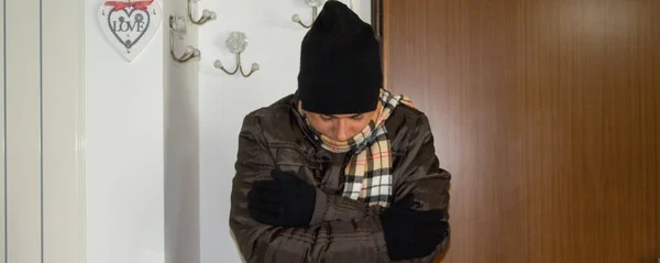 Image of a man at home who feels cold and is wearing a coat, scarf, gloves and wool hat. Reference to the high cost of gas and the constant increase in heating bills. Horizontal banner