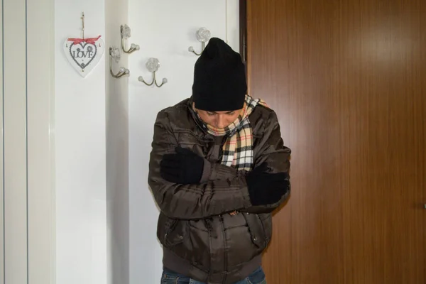 Image of a man at home who feels cold and is wearing a coat, scarf, gloves and wool hat. Reference to the high cost of gas and the constant increase in heating bills